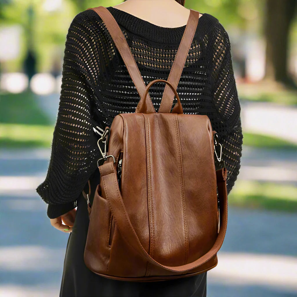 Person wearing a black knit sweater carrying a brown leather backpack from Luggo Bags, featuring an additional shoulder strap, while walking outdoors in a sunlit park.